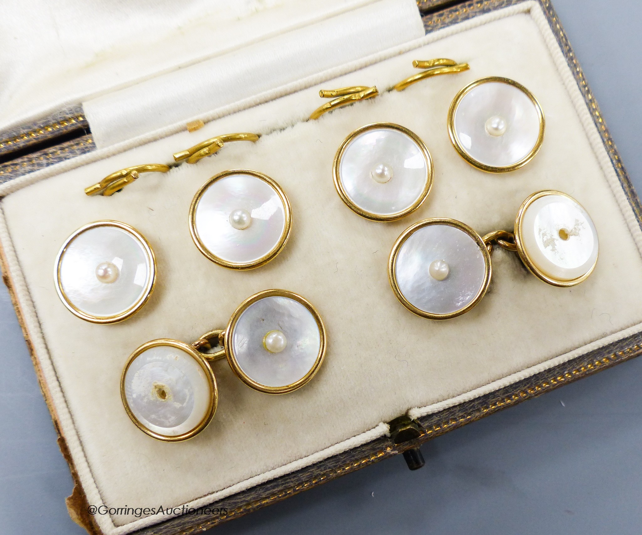 A cased six piece yellow metal (stamped 18), mother of pearl and seed pearl set circular dress stud set, (two pearls missing), gross 14.7 grams, in Goldsmiths & Silversmiths Co. Ltd.